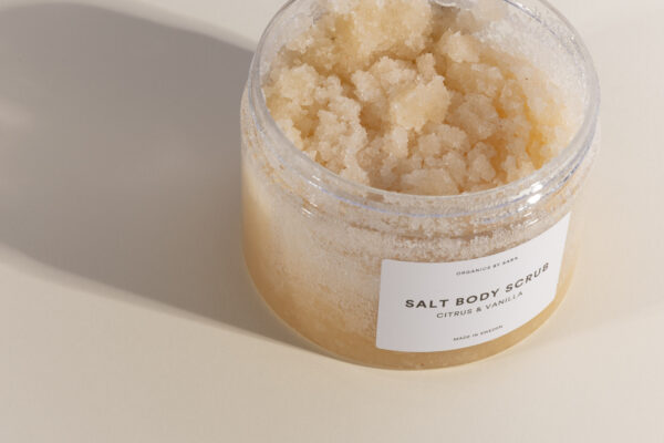 Burk med öppnat lock så att man ser inuti en aprikosfärgad salt scrub. Vit etikett på framsidan med Organics by Sara "Salt Body Scrub".